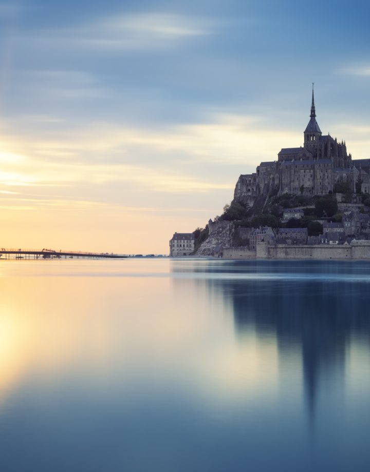 Le Mont-Saint-Michel au crépuscule, se reflétant les eaux calmes qui l'entourent pendant une grande marée, avec le soleil couchant créant des teintes dorées sur l'horizon.